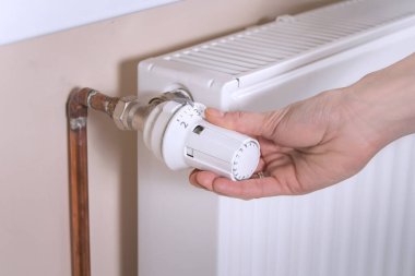 Kadının eli radyatör sıcaklık kontrol vanasını tutuyor ve gerekli değeri ayarlıyor. Beyaz radyatör ve bakır su boruları ikinci planda..