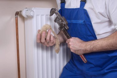 Bir muslukçu bir elinde ayarlanabilir anahtarı, diğer elinde de çekici tutar. Arka planda radyatör ve bakır tüplü bir duvar var..