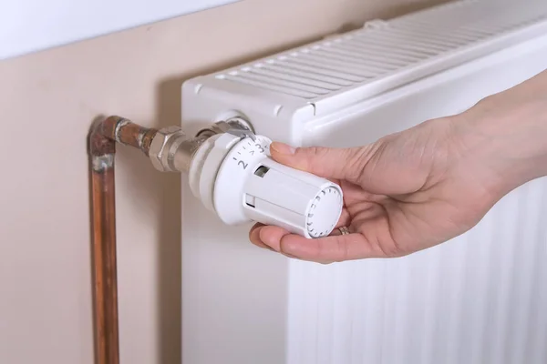 The woman\'s hand holds the radiator temperature control valve and sets the required value. White radiator and copper water pipes on the second background.