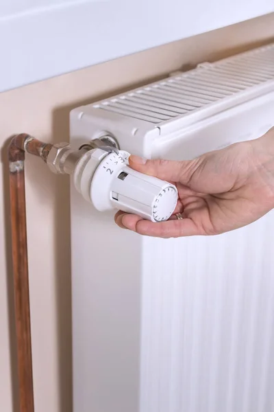 The woman\'s hand holds the radiator temperature control valve and sets the required value. White radiator and copper water pipes on the second background.