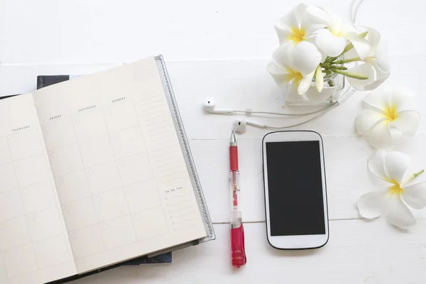 notebook planner  with mobile earphone for business and white flower frangipani on background white at office desk