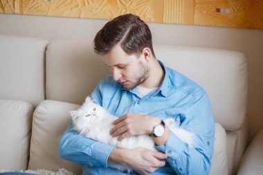 Mavi gömlekli beyaz kabarık kedi ile oynarken, çekici bir adam