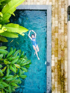 Genç bir kız bir havuzda sırtında palmiye ağaçları ile yatıyor. Yukarıdan kuşbakışı çekim.
