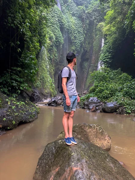 A young man stands on a rock in the middle of a river in the jungle. At the rear, the waterfall and he looks at the waterfall.