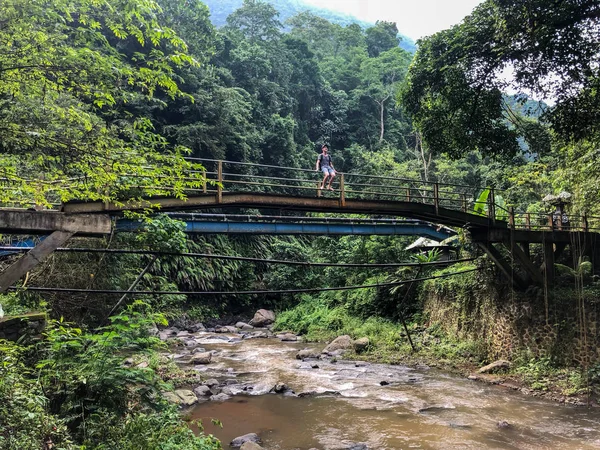 Bridge over the river in the jungle. A man is sitting on the bridge and watch. A shooting in the jungle on the island of Bali near the waterfall.