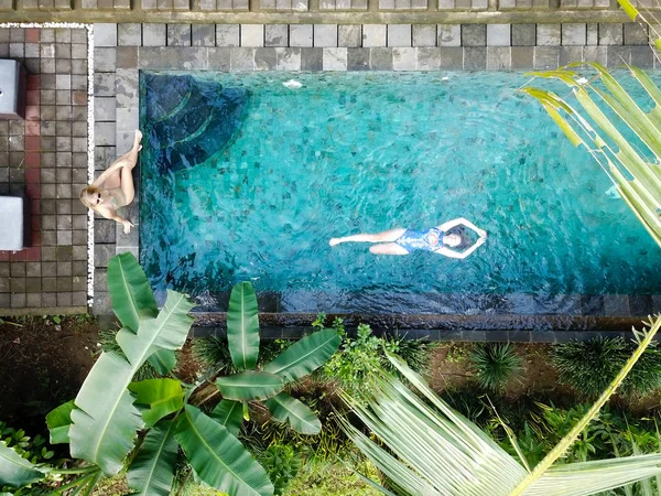 A young girl sits near the infinity pool with azure water. The second girl is swimming in the pool lying on her back. Villa in the jungle in Ubud on the island of Bali. Top view from the drone.