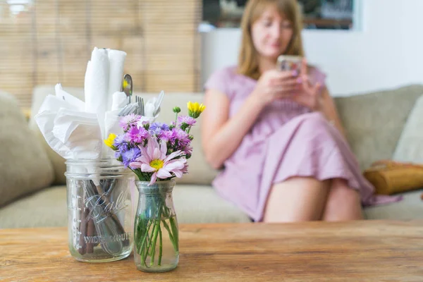 In the foreground there is a wooden table with a bouquet of flowers. On the back, blurry plan, the girl sits and looks into the phone.