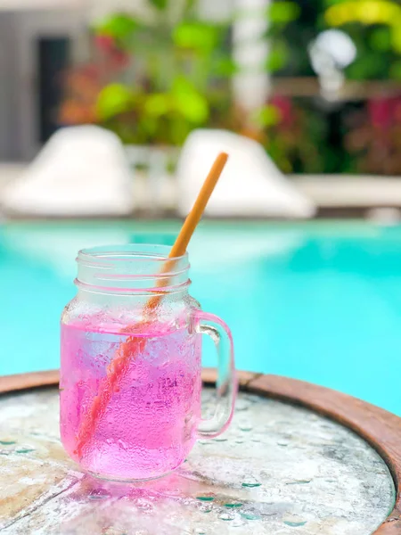 Pink drink - karkade in a transparent glass with a wooden straw on the background of the pool and on a white background. Side view with copy space