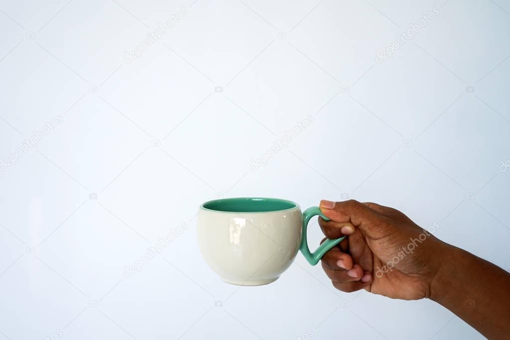 The hand of a dark-skinned man holds a white-green mug for coffee or tea against a white and blue wall. Side view with copy space