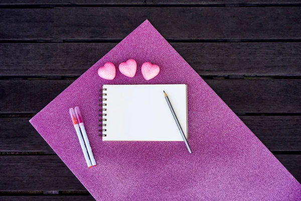 Minimal styled flat lay isolated on pink shiny and dark wood background. Girl desk top view with celebration accessories: heart, felt-tip pen, notebook, pencil. Woman's day postcard