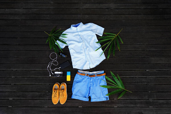 Man summer clothes collage flat lay isolated on dark wood background. Summer outfit of casual man desk top view fashion accessories: shirt, shorts, sunglasses, camera, smartphone. Holiday travel