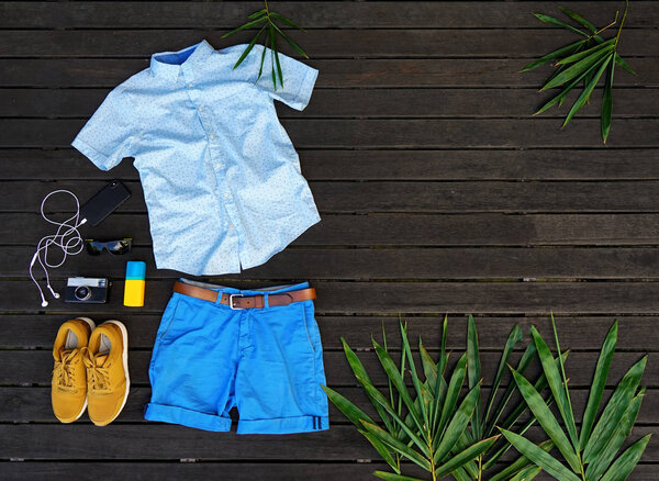 Man summer clothes collage flat lay isolated on dark wood background. Summer outfit of casual man desk top view fashion accessories: shirt, shorts, sunglasses, camera, smartphone. Holiday travel