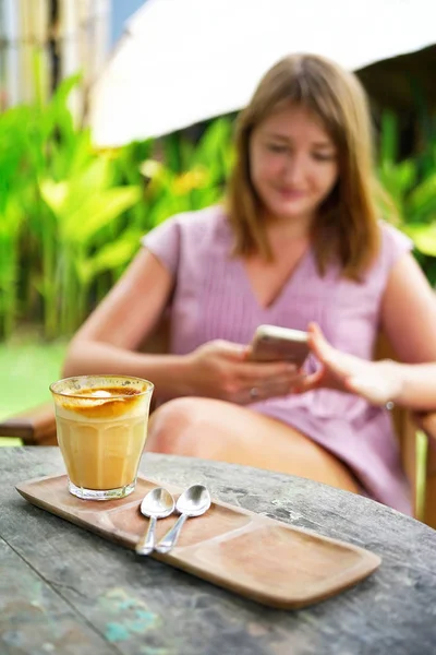 A young red-haired girl with a pastel dress is sitting at a table in a cafe and looking into her smartphone. On the table is breakfast, coffee, water. Side view background is blurred