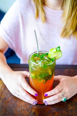 Genç bir kız bir kafede ahşap bir masada otururken bir serinletici turuncu kokteyl içer. Yaz, güneş, iyi ruh hali. Çerçeve close-up modeli eller. Kopya alanı ile yan görünüm