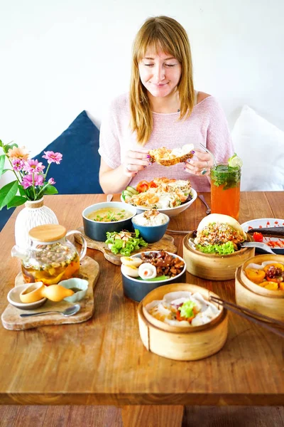 A large, wooden table. A young, beautiful girl in pink is sitting at the table. On the table there are many different dishes of French and Chinese cuisine: salads, dumpling, meat stew and many other