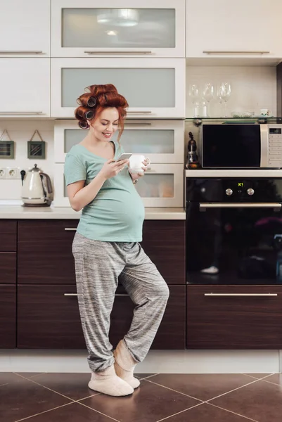 Gravid kvinna använder en smartphone stående i köket — Stockfoto