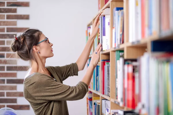 Молодая женщина в очках ищет книгу на книжной полке — стоковое фото