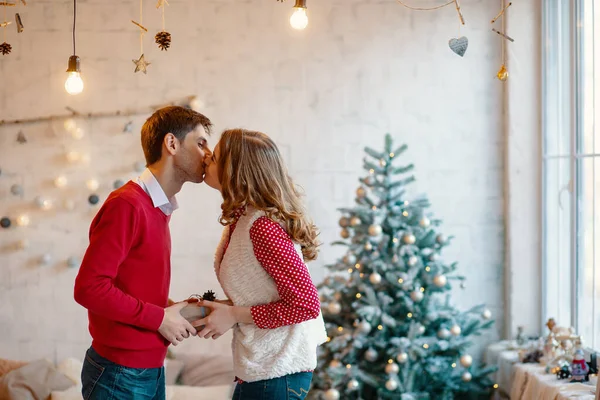 Junges Paar küsst sich beim Zusammenhalten einer Geschenkschachtel, mit Weihnachtsdekoration im Hintergrund — Stockfoto