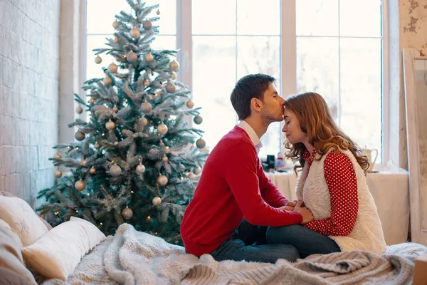 Glückliches Liebespaar, das Weihnachten genießt. junger Mann küsst seine Freundin auf die Stirn, berührt ihre Hände, sitzt auf einem Bett mit geschmücktem Baum und Fenster im Hintergrund — Stockfoto