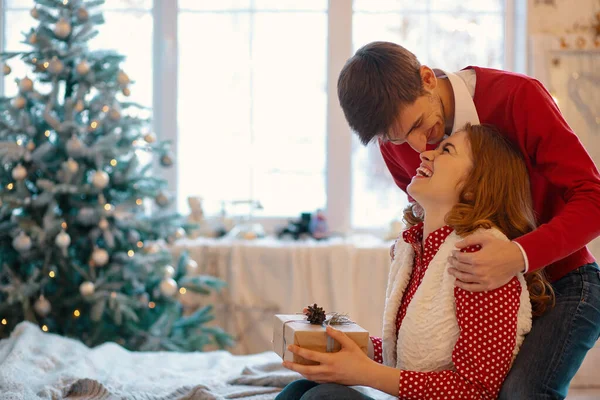 Glückliches Liebespaar, das Weihnachten genießt. junger Mann berührt seine Freundin mit der Nase und hält ein Geschenk mit Fenster im Hintergrund — Stockfoto