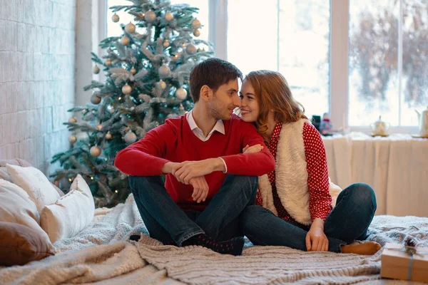 Junger Mann, der lächelnd in die Augen seiner Freundin blickt, sitzt mit überkreuzten Beinen auf einem Bett mit geschmücktem Baum und Fenster im Hintergrund. glückliches Liebespaar genießt Weihnachten. — Stockfoto
