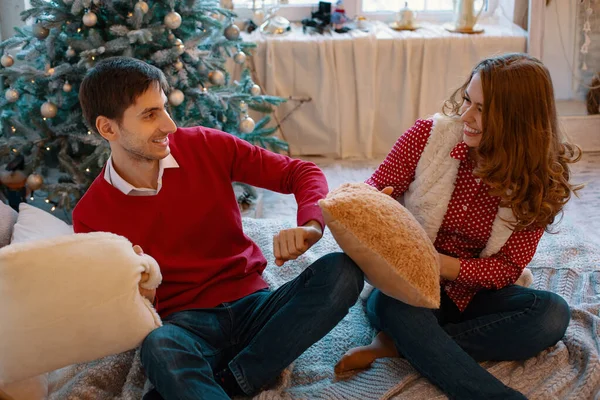 Glücklich liebendes kaukasisches Paar wird an Heiligabend in der Nähe des Neujahrsbaums verrückt, sie sitzen auf dem Bett, lachen viel, kämpfen mit Kissen — Stockfoto