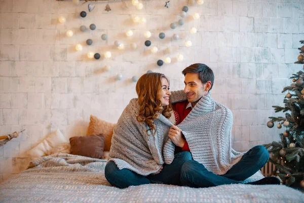 Junger Mann, der lächelnd in die Augen seiner Freundin blickt, sitzt mit überkreuzten Beinen auf einem Bett mit geschmücktem Baum und Fenster im Hintergrund. glückliches Liebespaar genießt Weihnachten. — Stockfoto
