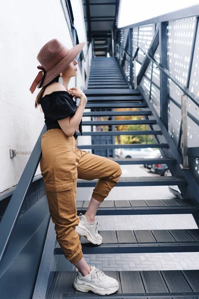 Divatos barna nő hosszú hajjal, elegáns nagy fonott kalapot visel, pózol a lépcsőn — Stock Fotó