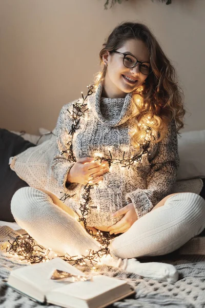 Junge zukünftige Mutter sendet positive Vibes an ihr ungeborenes Kind, indem sie ihre Kleidung mit Lichtern dekoriert, während sie es sich zu Hause auf dem Bett gemütlich macht — Stockfoto