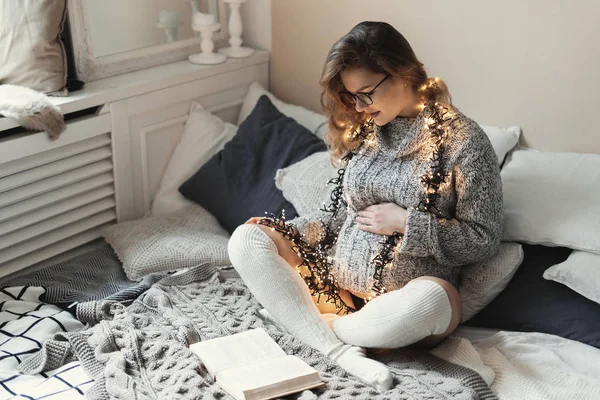 Young future mom sending positive vibes to her unborn child by decorating her clothes with lights while relaxing on bed at home — 스톡 사진