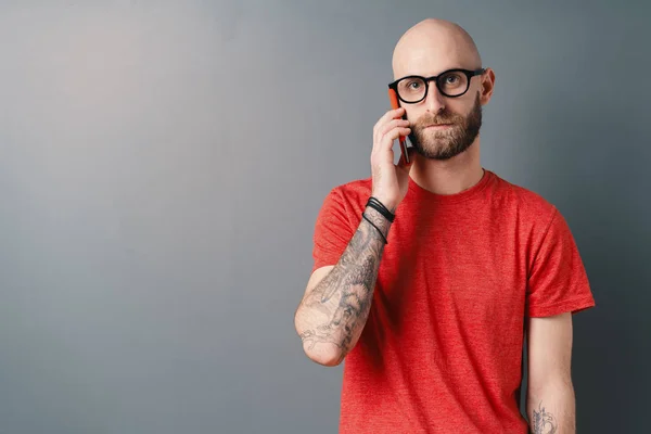 Красивый белый мужчина без волос с бородой, в очках, в красной футболке с телефоном на фоне серой студии — стоковое фото