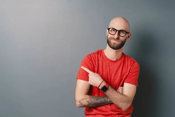 Stílusos fiatal szakállas férfi rögzíti a fekete keretes szemüveg egy tetovált kézzel, miközben mosolyog a stúdióban — Stock Fotó