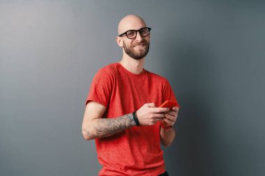 Tüysüz, sakallı, gözlüklü, kırmızı tişörtlü, akıllı telefonu dövmeli kollarında tutarken gri stüdyo arka planında duran yakışıklı beyaz adam..