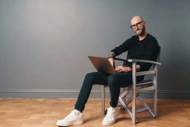 Stüdyonun içinde sakallı ve gözlüklü gülümseyen beyaz adam gümüş koltukta oturuyor, gri arka planda oturuyor ve bilgisayarda internette sörf yapıyor. Cihaz kullanarak online iletişimden hoşlanan adam.