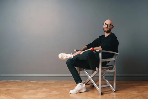 A modern, stylish man sitting leg over leg, in a confident pose on a silver chair in the studio on a gray wall background. He wears a beard, glasses, black casual clothes and white sneakers.