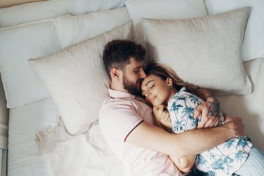 Aşık çift yatakta kucaklıyor. Adam kız arkadaşına sarılıp öpüyor. Üstte görüntü