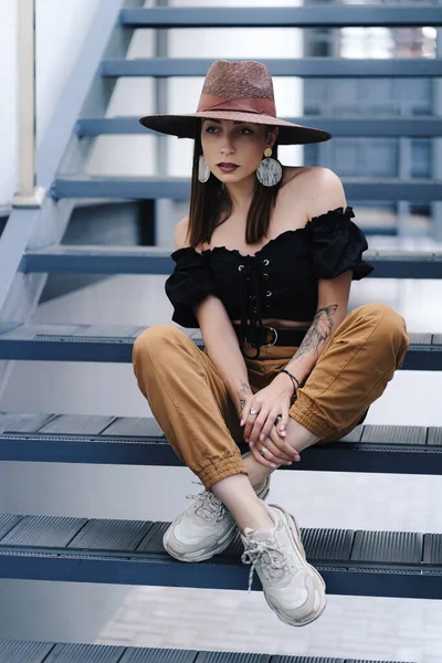 Mulher morena elegante com cabelo longo, vestindo chapéu de vime grande elegante, posando nas escadas — Fotografia de Stock