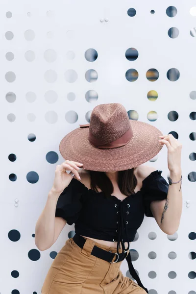 Mujer morena de moda con el pelo largo, con elegante sombrero de mimbre grande, posando contra una pared blanca con agujeros —  Fotos de Stock