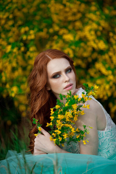 Beautiful young girl with red hair in a lavish dress walking in the nature with flowers