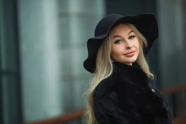 年轻美丽的女孩在黑色毛皮大衣和帽子在街道附近的建筑物 — 图库照片