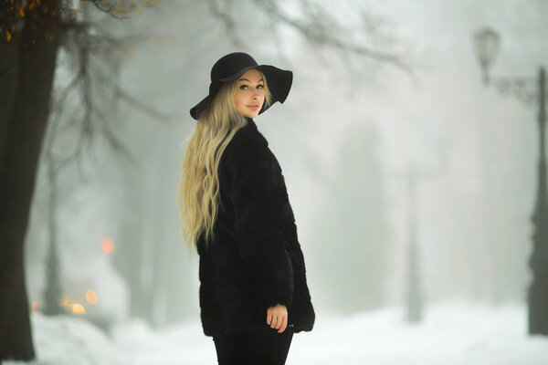 Beautiful young girl in fur coat and hat walking in the park in winter weather