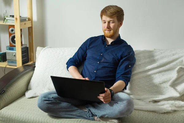 handsome young guy with a beard working at home with a laptop sitting on the couch