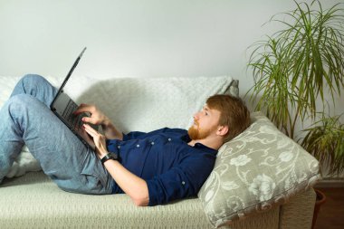 evde kanepede yatan bir dizüstü bilgisayar ile çalışan bir sakallı yakışıklı genç adam