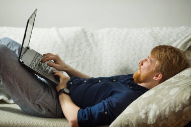 evde kanepede yatan bir dizüstü bilgisayar ile çalışan bir sakallı yakışıklı genç adam