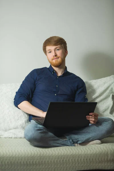 handsome young guy with a beard working at home with a laptop sitting on the couch