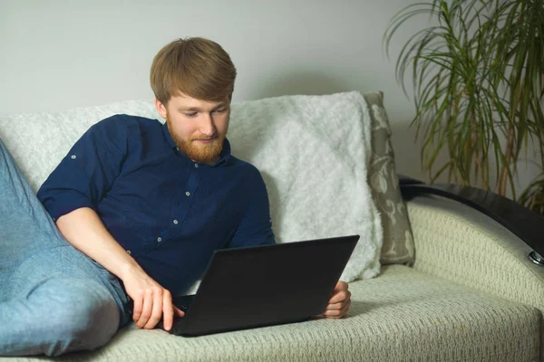 handsome young guy with a beard working at home with a laptop lying on the couch