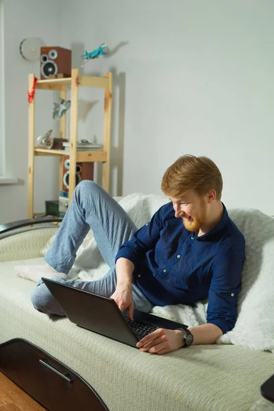 handsome young guy with a beard working at home with a laptop lying on the couch