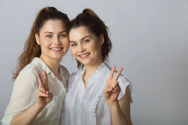 两个美丽的微笑的女孩姐妹双胞胎在白色上衣在光的背景显示手势成功 — 图库照片