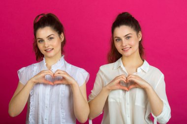 iyi bir ruh hali içinde bir el gösterilen pembe bir arka plan üzerinde beyaz bluz iki güzel genç kız ikiz bir kalp şeklinde jest