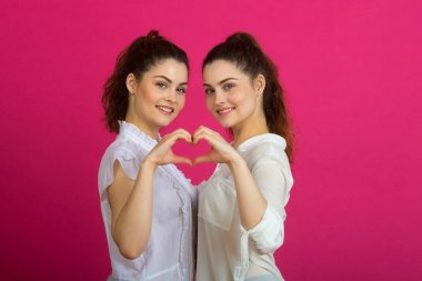 iyi bir ruh hali içinde bir el gösterilen pembe bir arka plan üzerinde beyaz bluz iki güzel genç kız ikiz bir kalp şeklinde jest